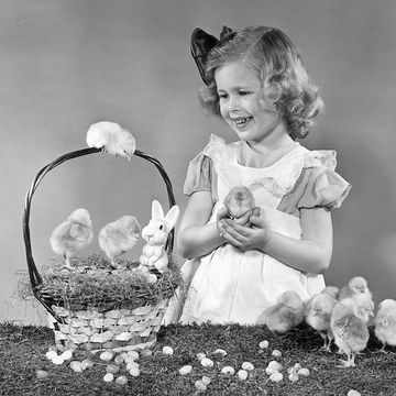 black and white photo of young girl holding a baby chick and standing next to an easter basket