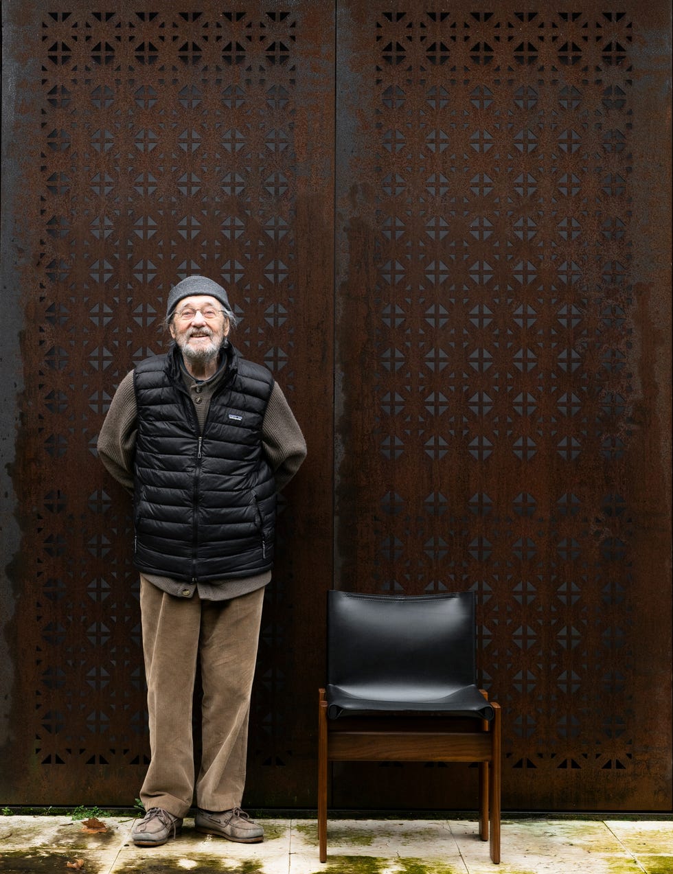 A person stands against a patterned metal wall with a chair beside them.