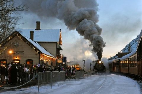 antique train with smoke billowing from it's stack pulling into an old fashioned depot with people lined up waiting to get on the train it is dusk out, and there is snow on the ground