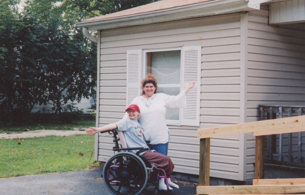 gypsy rose blanchard sits in a wheelchair with her mother standing next to her, they smile in front of a house with a wheelchair ramp and each reach one arm out to the side