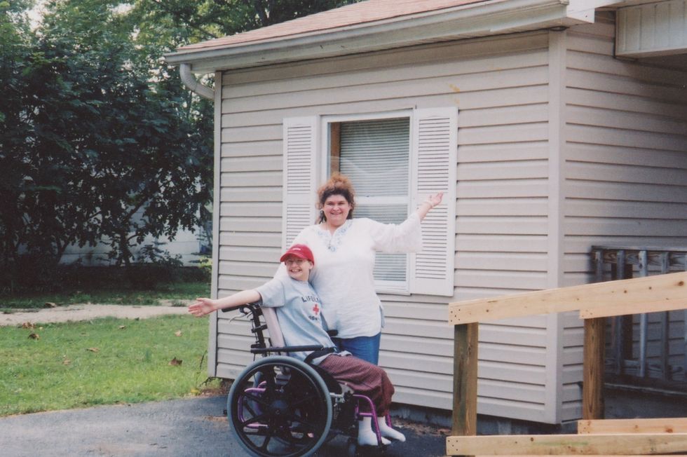 gypsy rose blanchard sits in a wheelchair with her mother standing next to her, they smile in front of a house with a wheelchair ramp and each reach one arm out to the side