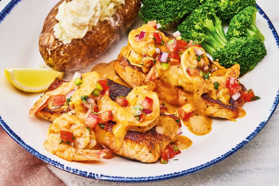 plated dish featuring seasoned fish topped with shrimp and sauce alongside a baked potato and broccoli