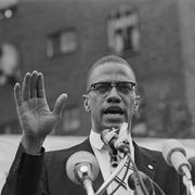 malcolm x speaks into several microphones, he has his right hand raised in front of him, he wears a suit and his signature glasses