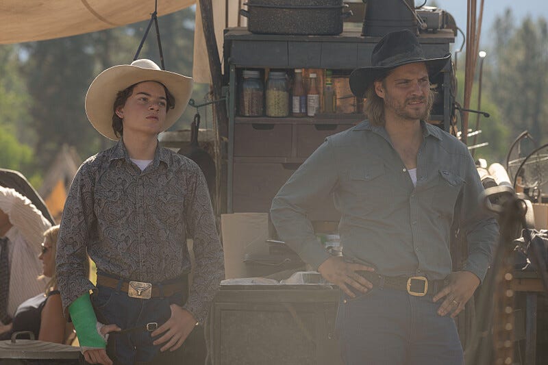 Two individuals in western attire standing in front of a wagon filled with supplies