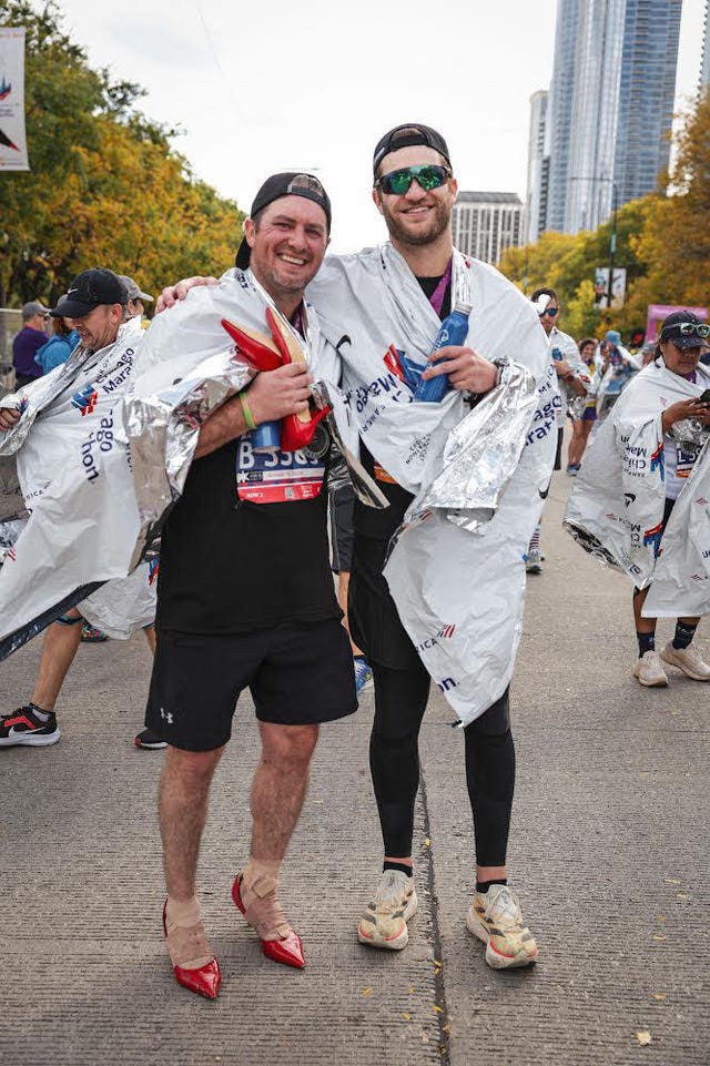 two individuals stand on a street post marathon, one wearing red high heeled shoes and the other in athletic gear both are wrapped in metallic recovery blankets in the background, other participants can be seen, along with a city skyline featuring tall buildings and colorful trees