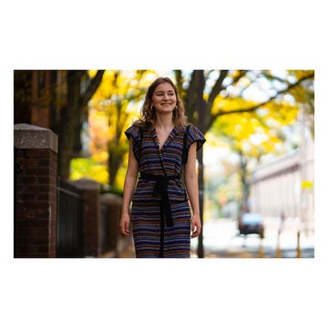 a woman stands confidently on a street lined with trees, showcasing a dress that features a colorful geometric pattern and a belt tied at the waist the background displays a blurred view of a street with foliage showing autumn colors, enhancing the outdoor ambiance