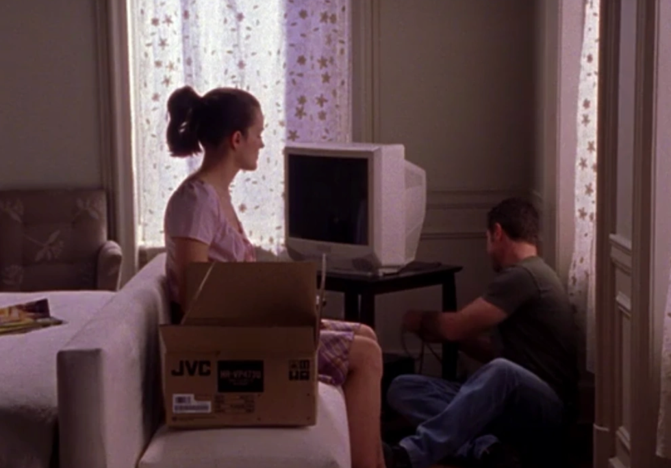 A person sits on a couch beside a box while another person connects wires near a television.
