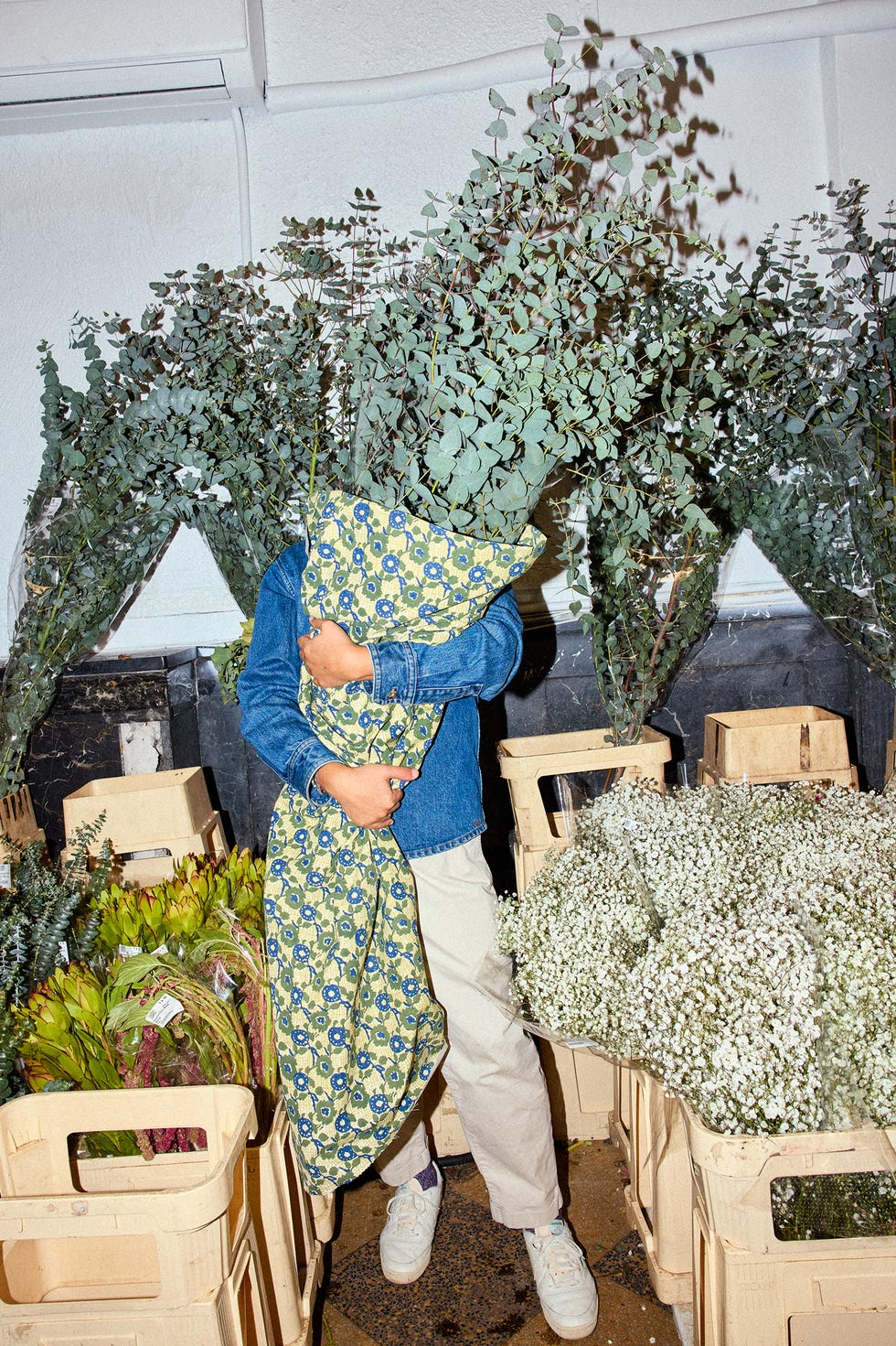Person holding eucalyptus branches while wrapped in a patterned fabric, surrounded by floral arrangements.