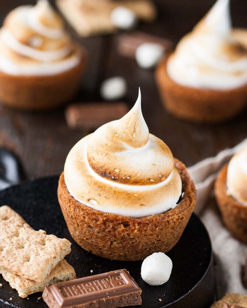 s'mores cookie cups with a cookie cup crust, chocolate filling, and toasted meringue topping, on a plate with graham crackers, hershey's chocolate square, and marshmallow
