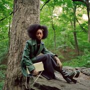 model sits on rick in the woods wearing a green blazer and carrying a bag