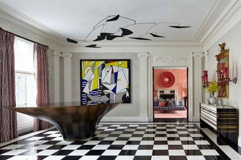 Large open plan foyer with dramatic black and white floor tiles and a large brown oblong pedestal table in one corner and behind it a large Picasso-like modern painting in white and black with touches of yellow and blue and opposite the table a striped console with red Murano glass like candelabra above it on the wall and through the door you can see the pink living room