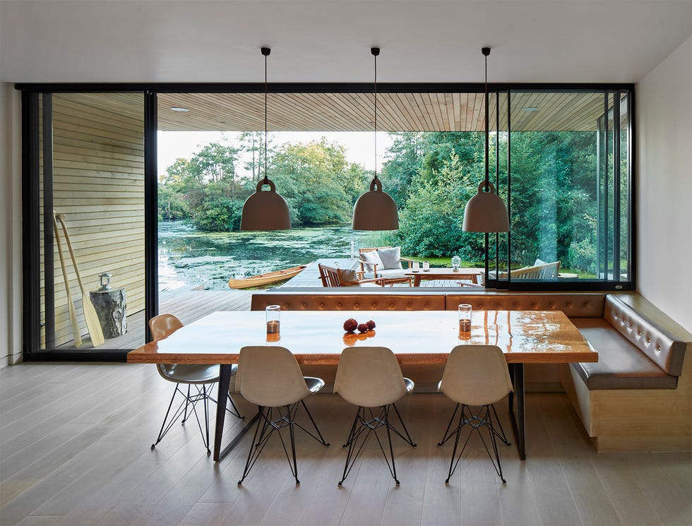 a wooden dining table with chairs and a banquette, framed by large sliding glass doors that look out to a lagoon with greenery