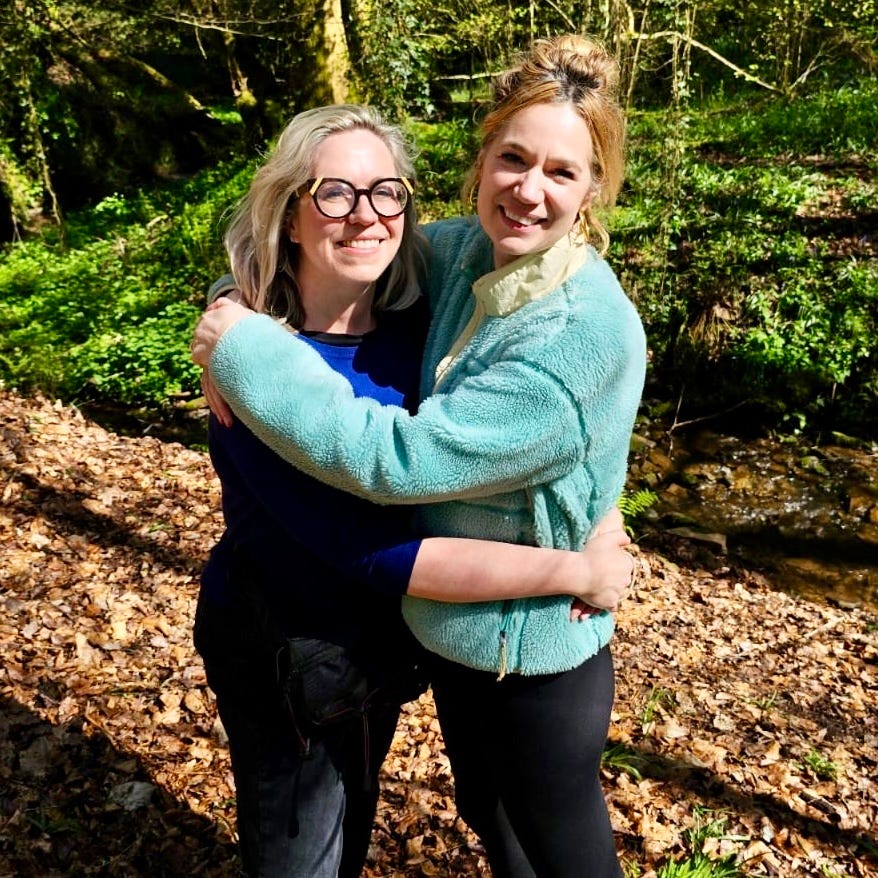 two women hugging in a wooded area with sunlight filtering through the trees