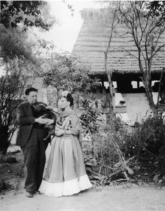 Frida Kahlo and Diego Rivera stand together with a pet dog in front of thatchted-roof hut which houses a number of archeological artifacts, Mexico City, Mexico