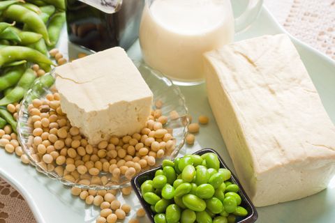 loose soy beans, tofu, and soy milk, full block of tofu  half block of tofu sitting on plate of loose soy beans green beans in black square bowl glass filled with soy milk