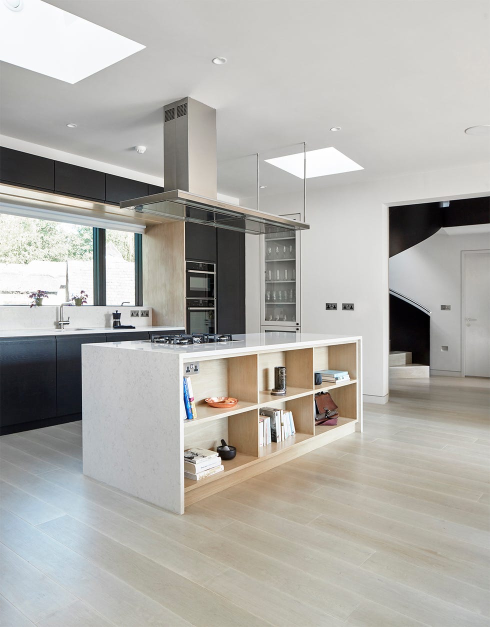 a white washed kitchen with a stone island, black cabinetry and pale wooden floors, with light pouring in from a picture window and two velux roof windows