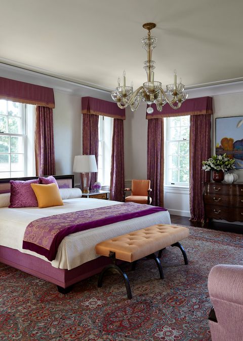 Bedroom with floor to ceiling cranberry toned curtains, patterned carpet and throw on the bed and a small light persimmon tufted bench with modern black legs in front of the bed