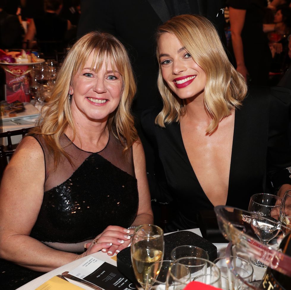 tonya harding and margot robbie smile at the camera while sitting at a table filled with glasses and other items, both women wear black dresses and have shoulder length blonde hair