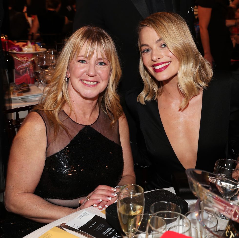 tonya harding and margot robbie smile at the camera while sitting at a table filled with glasses and other items, both women wear black dresses and have shoulder length blonde hair