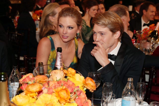 taylor swift and joe alwyn sit at a table with yellow and orange flowers and several drinks on it, she wears a sleeveless blue and orange dress, he wears a black tuxedo and bowtie, they look to the left