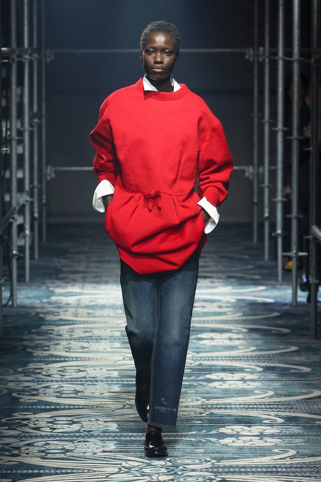 Un mannequin montrant un pull rouge surdimensionné et un pantalon en jean bleu sur la piste.