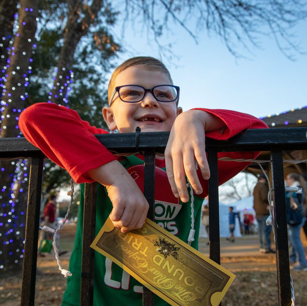 little smiling boy in glasses draped over a railing holding a gold colored polar express ticket there are trees lit in blue bulbs behind him