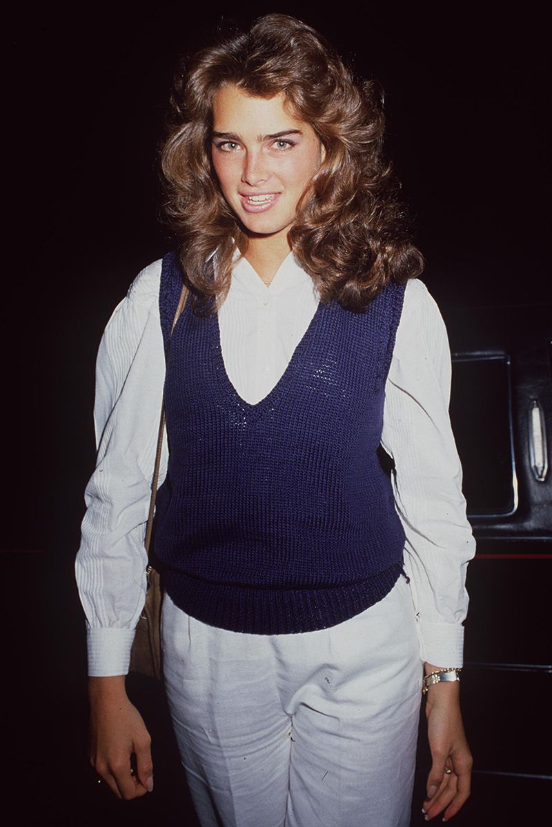 Brooke Shields wearing a white blouse and trousers, finished with a blue sweater vest over the top and voluminous hair.