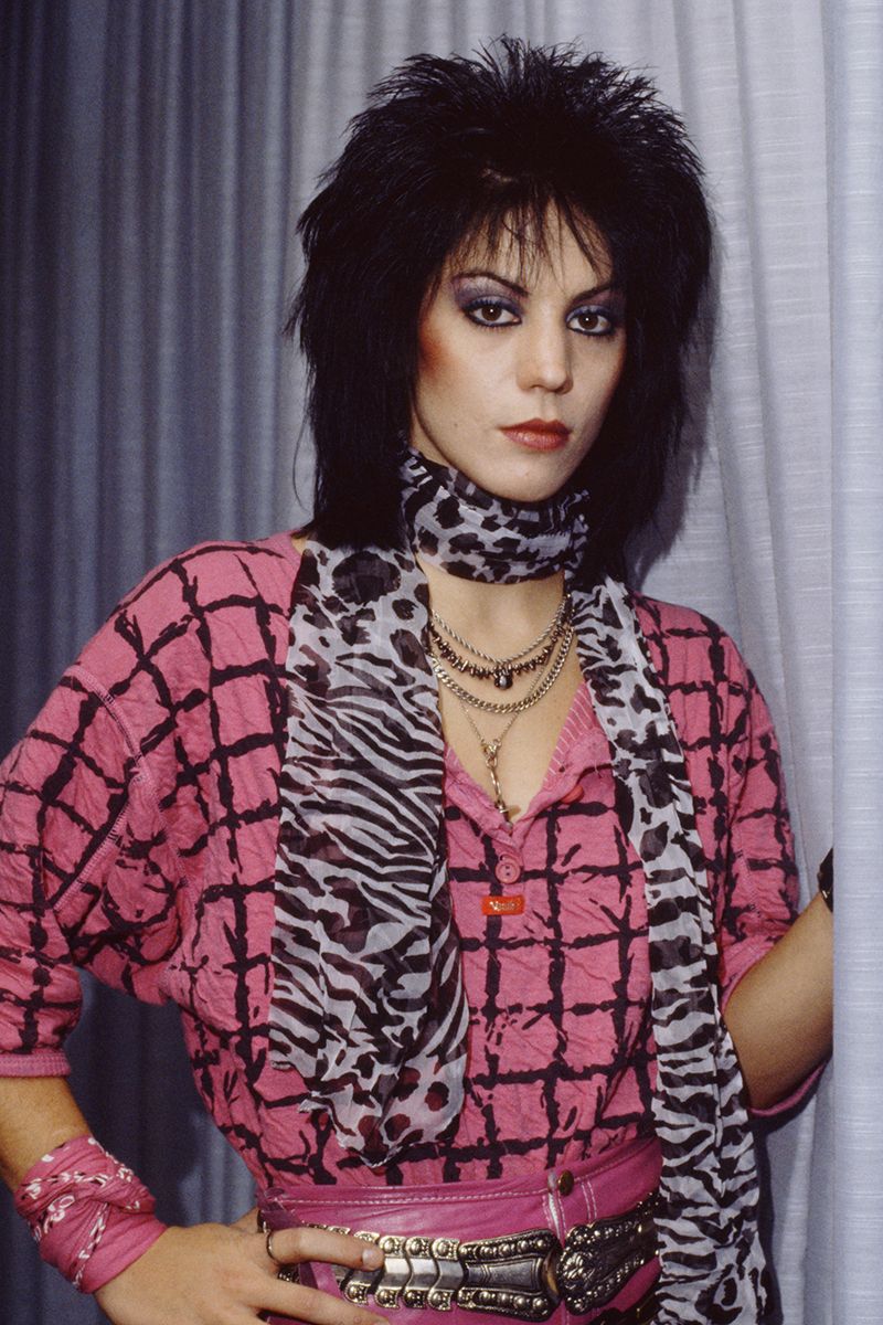 Joan Jett standing backstage wearing a pink check print top, zebra scarf, bandana bracelets, big hair, dark contouring and heavy eye makeup