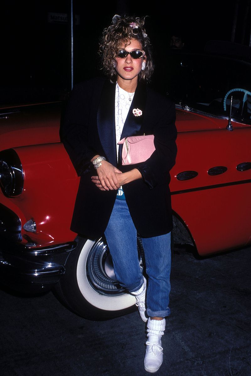 Sarah Jessica Parker in the 80s wearing mum jeans, a white top, black blazer and white trainers while leaning against a red car. The star finishes the outfit with a red lip, a messy up-do hair style and big earrings.