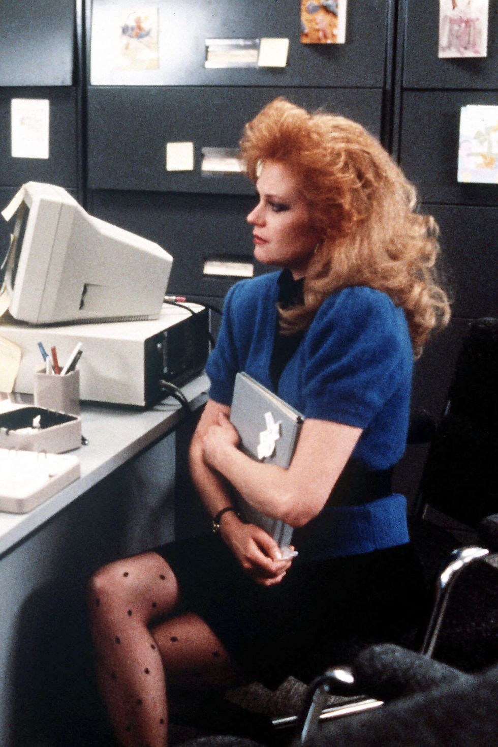 Melanie Griffith in Working Girl sitting in an office wearing a blue cardigan over a black dress with polka dot tights and big hair