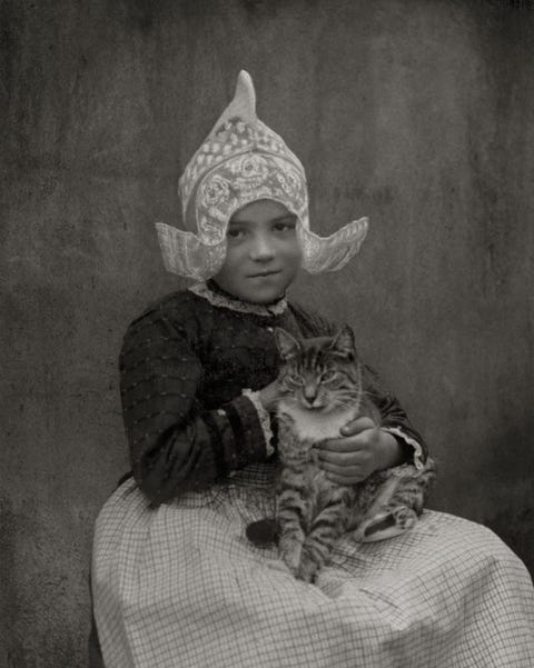 Dit meisje uit het Volendam van de jaren twintig heeft haar kat op schoot terwijl ze wat argwanend naar de fotograaf kijkt