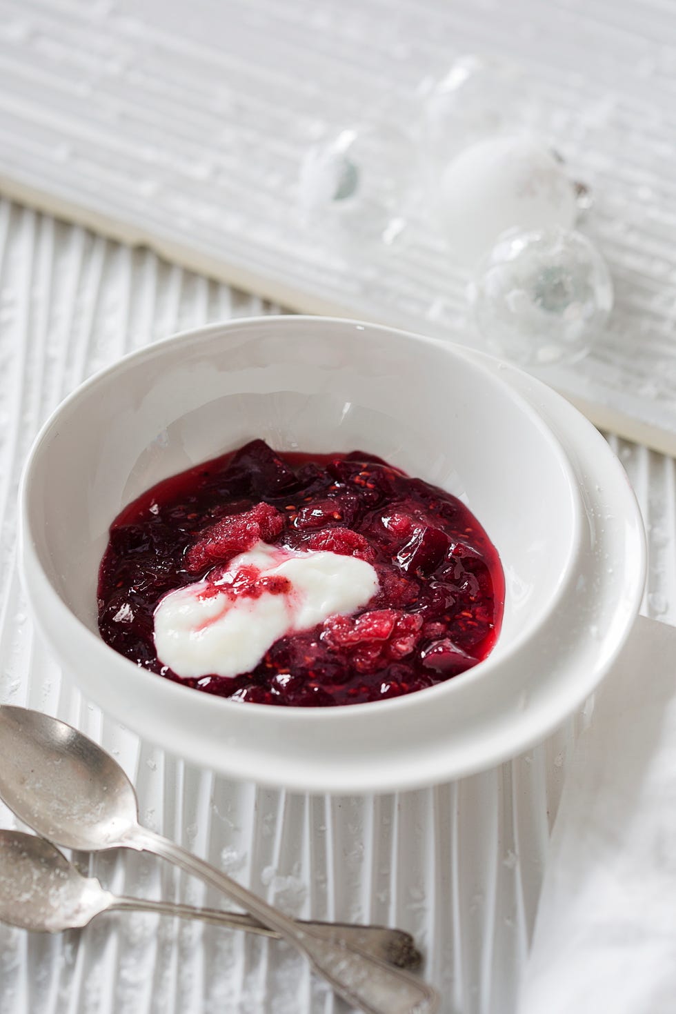 bowl of raspberry sauce with a dollop of cream surrounded by utensils and a soft white surface