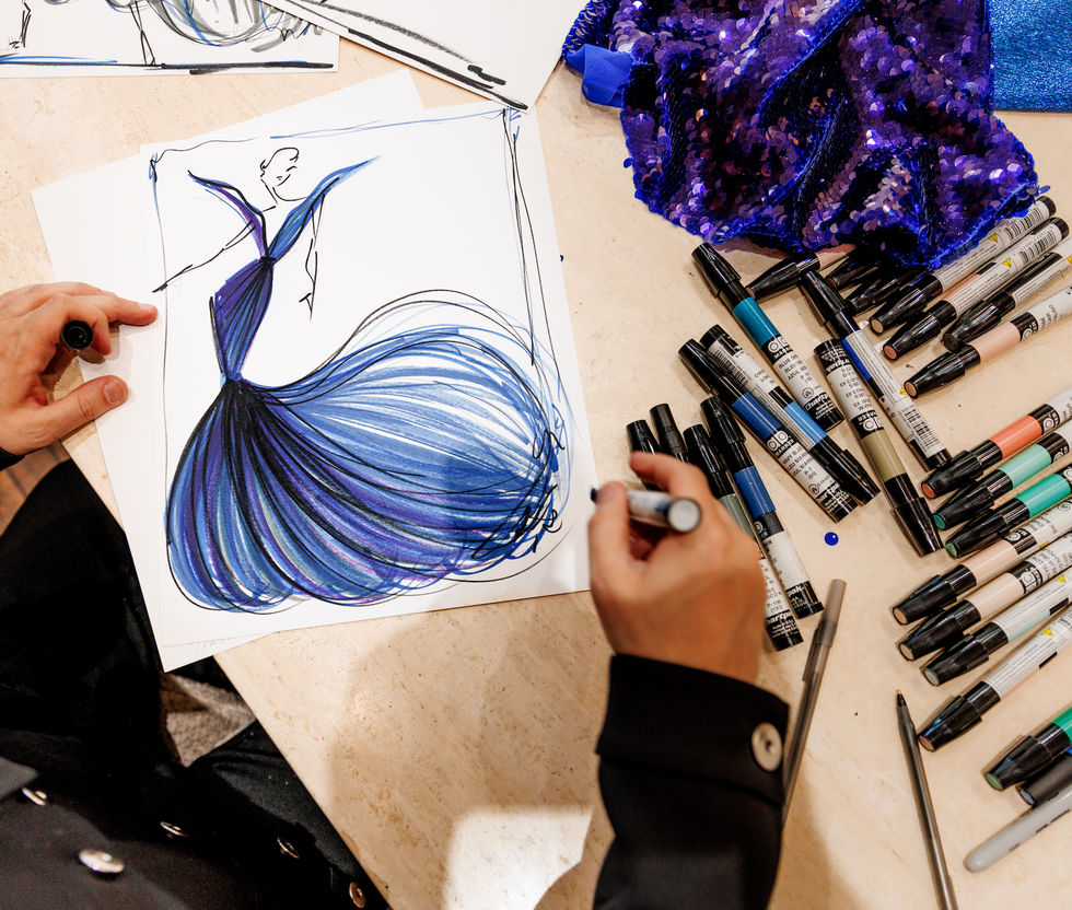 Fashion designer sketching a gown on paper with various markers nearby.