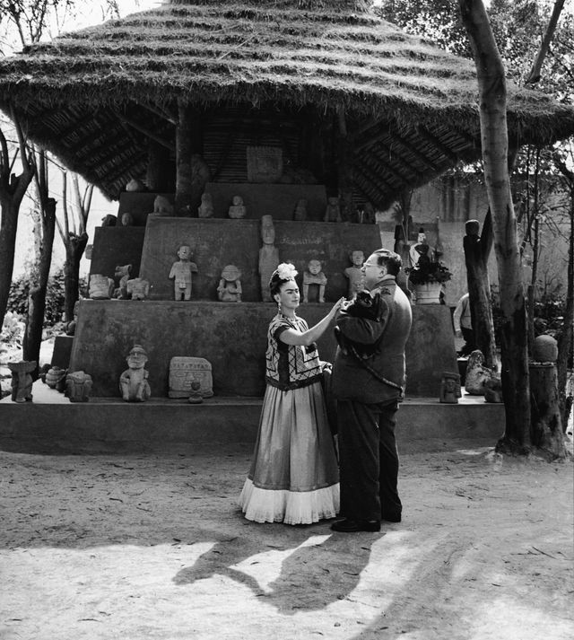 Frida Kahlo and Diego Rivera stand together with a pet monkey in front of thatchted-roof hut which houses a number of archeological artifacts, Mexico City, Mexico,