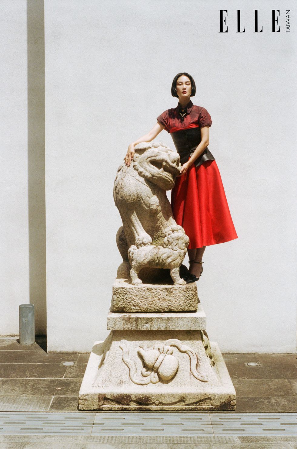 短髮女生身穿紮染襯衫、紅色平口洋裝、皮革馬甲，站立於石獅雕像旁。