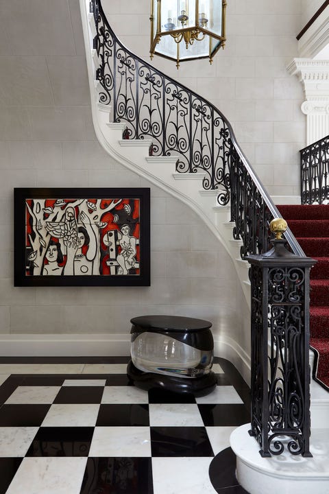another view of the foyer with black and white tiles and a dramatic white staircase with black banisters with a gold ball knob and crimson runner and on the wall below is another modern piece of art in red, white and black and a small oval mercury glass table with Black base and a black top