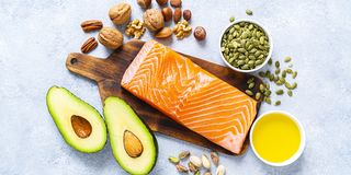 overhead view of a group of food rich in healthy fats the photo includes salmon, avocado, extra virgin olive oil, nuts and seeds like walnut, almonds, pecan, hazelnuts, pistachio and pumpkin seeds
