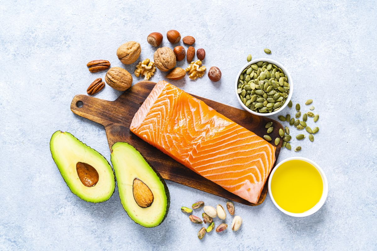 overhead view of a group of food rich in healthy fats the photo includes salmon, avocado, extra virgin olive oil, nuts and seeds like walnut, almonds, pecan, hazelnuts, pistachio and pumpkin seeds