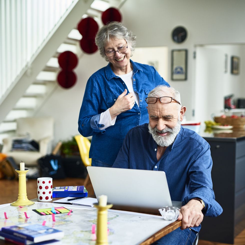 senior woman smiling and looking over husband's shoulder as he researches and reads online reviews about places to stay, and locations to visit, sticky notes and guidebooks on table, booking holiday online
