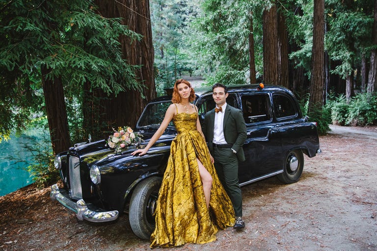 a man and woman standing next to a car in a wooded area