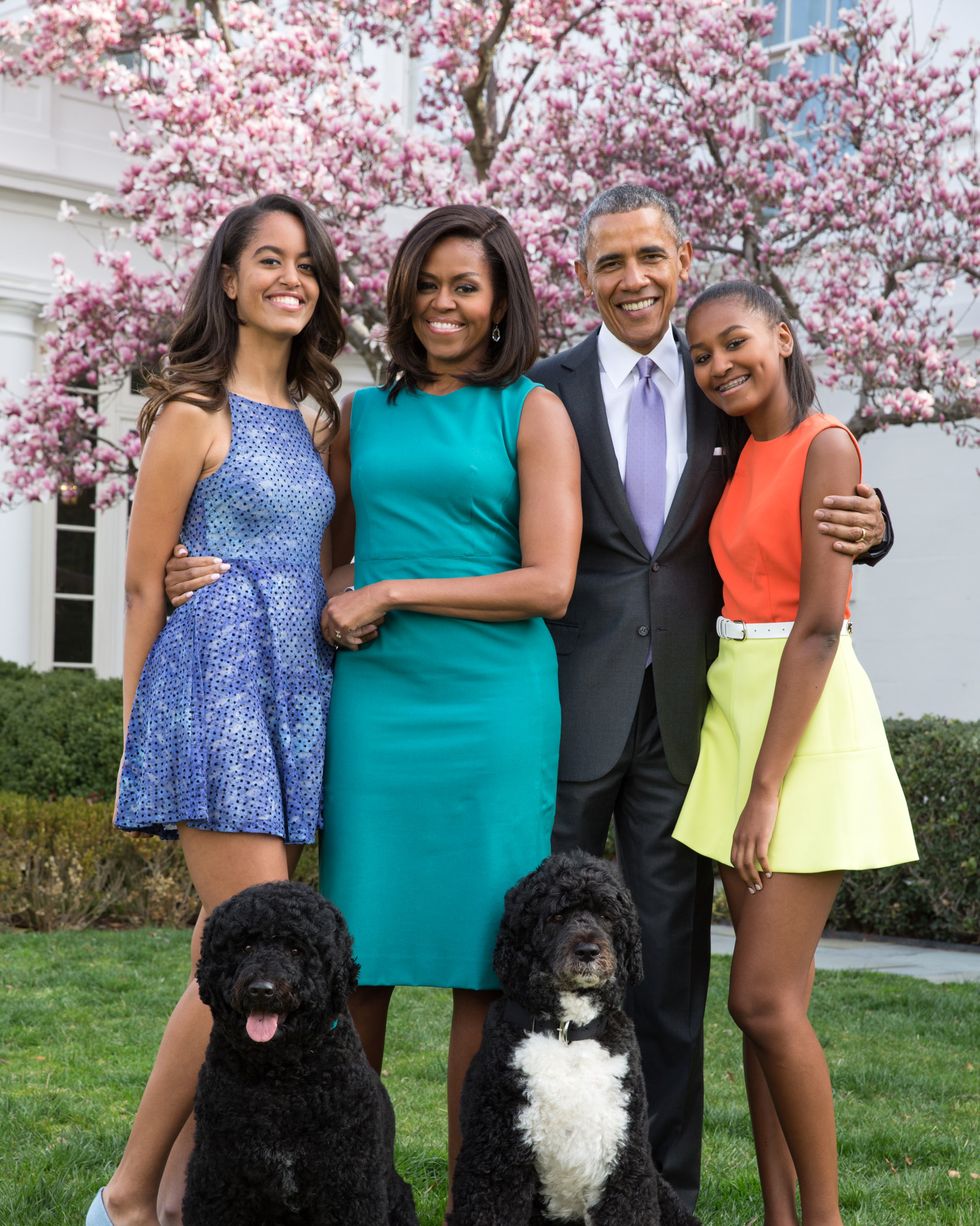 malia obama, michelle obama, barack obama, and sasha obama smile at the camera while standing on a lawn outside the white house, sitting in front of them are their two dogs, all four family members are wearing formal attire