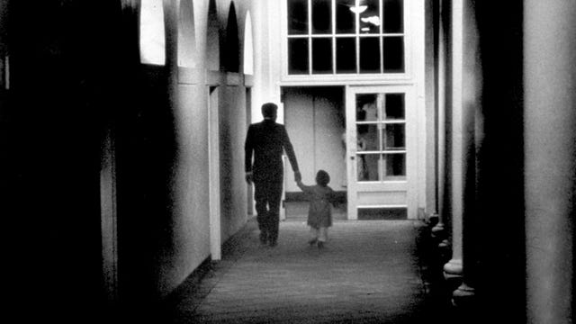 John F, Kennedy walking along a dark White House colonnade with his toddler son, John-John, seen from behind.