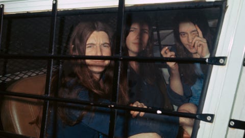 three female members of the manson family sit inside a police vehicle with cars on the window