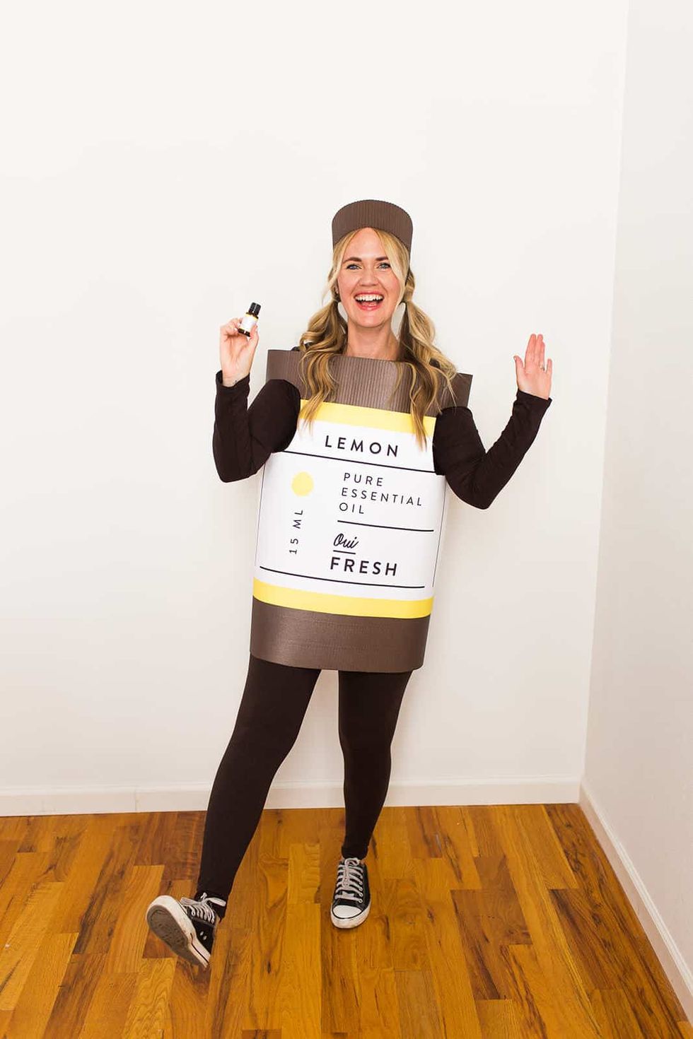 woman in black pants and long sleeved black shirt and a brown pillbox hat dressed as a bottle of essential oil that is made of of brown corrugated cardboard with a white label that reads lemon pure essential oil fresh she is holding a small bottle of essential oil up