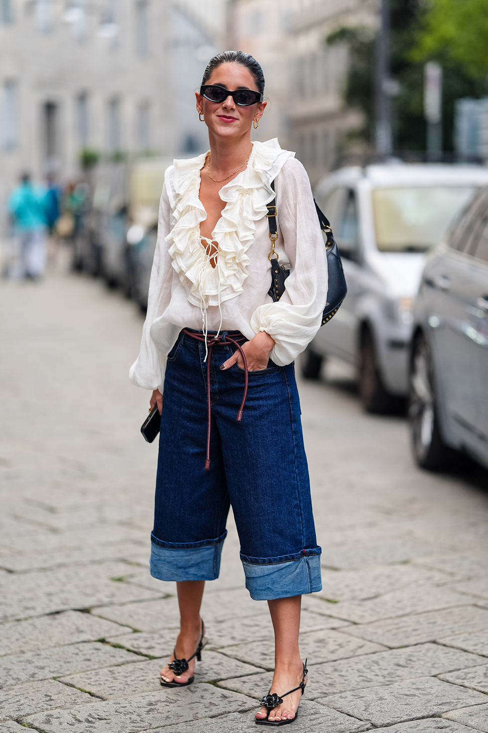 Milan, Italie, 20 septembre : une invitée porte des lunettes de soleil noires, des boucles d'oreilles dorées, une chemise blanche à manches longues à volants, un sac en cuir noir, un jean bleu marine foncé à plis, des tongs noires et des chaussures en cuir à détails floraux, devant Sportmax, lors de la Fashion Week de Milan printemps/été 2024-2025, le 20 septembre 2024 à Milan, en Italie. (Photo par Edward Berthelot/Getty Images)
