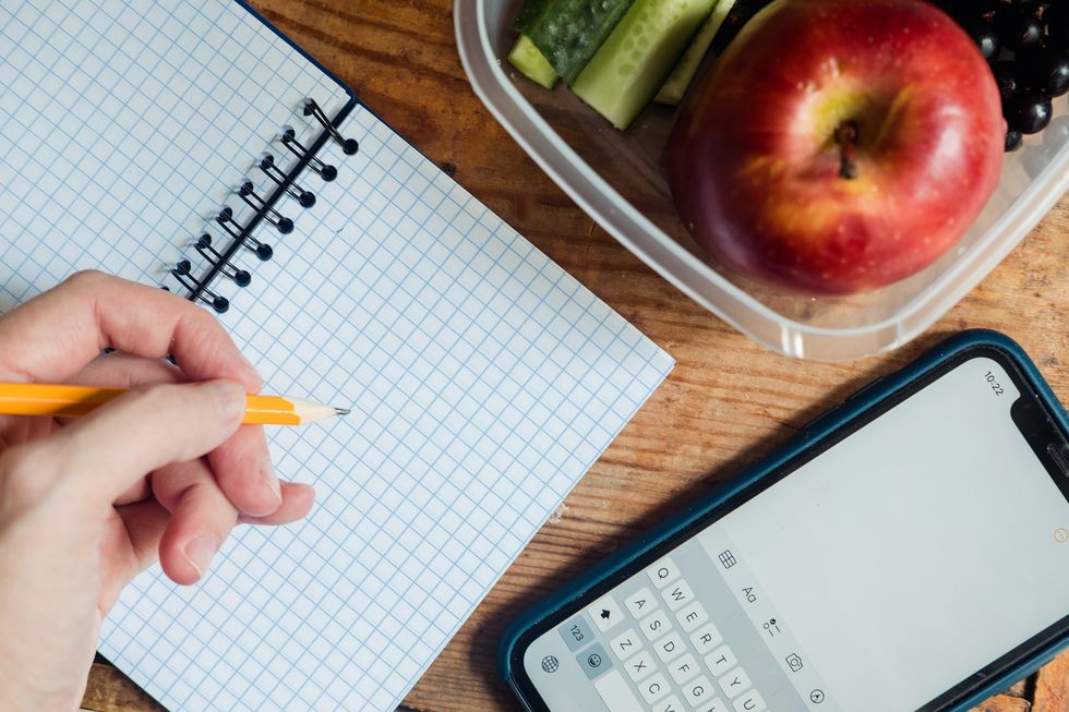 a hand with a simple pencil writes on a blank sheet of a notebook, during a break, next to the phone and a lunchbox with a red apple and cucumbers on a wooden background, top view