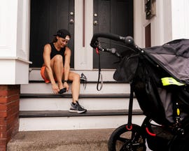 running postpartum woman sits on steps and puts her running shoes on while a stroller sits on the sidewalk