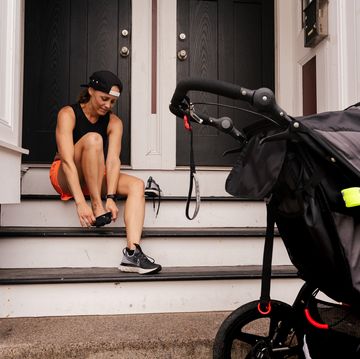 running postpartum woman sits on steps and puts her running shoes on while a stroller sits on the sidewalk