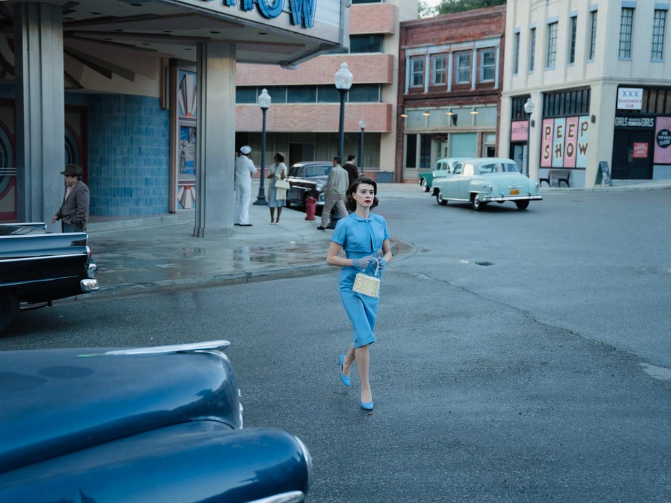 a woman in a vintage blue dress walks down a street with classic cars and historic buildings