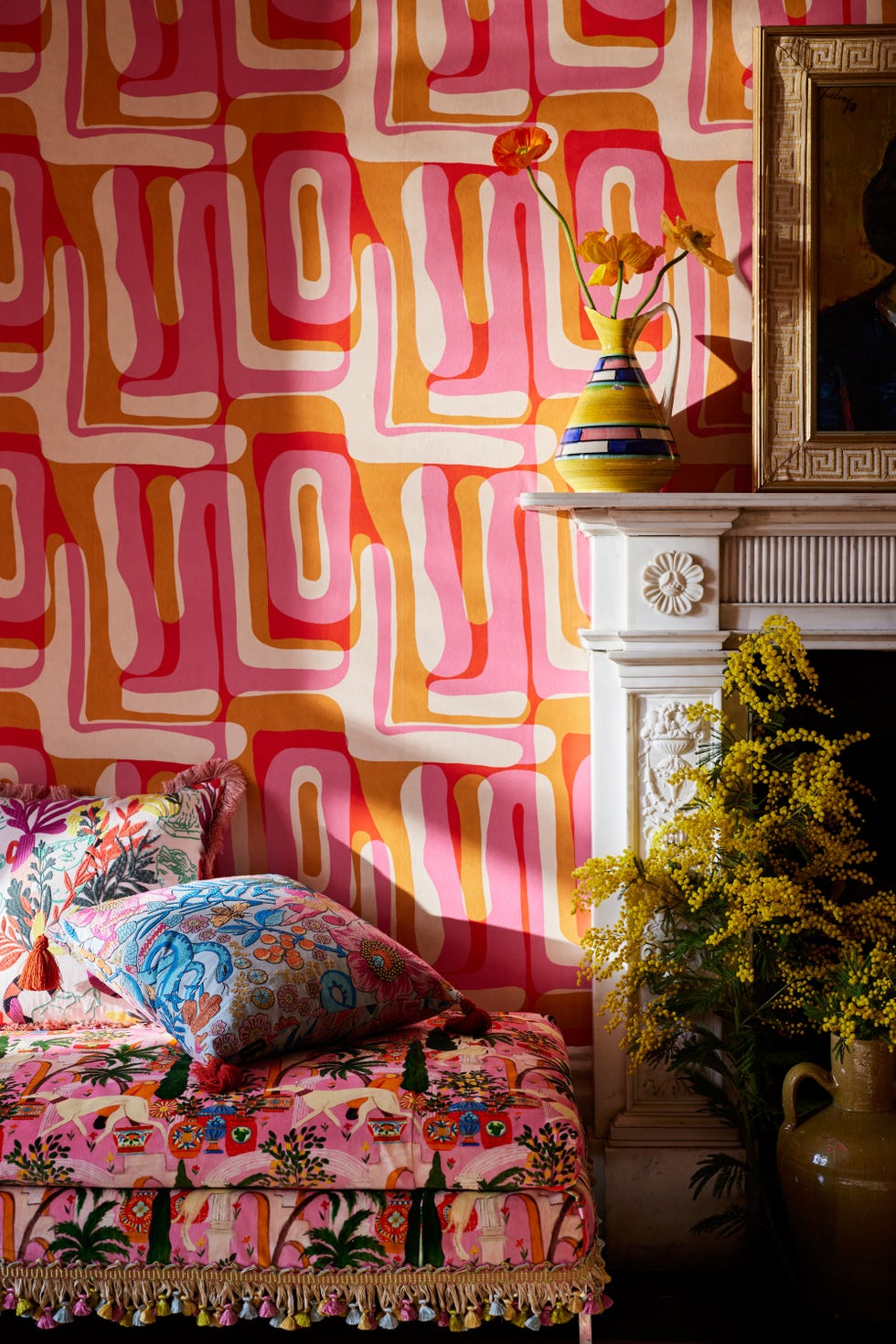 a room with pink, cream, and orange retro wallpaper, a pink floral bench with cushions on it, and a white fireplace with a vase and photo frame on it